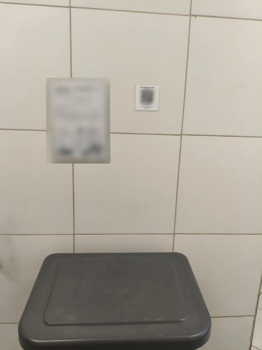 Banheiro com etiqueta de QR Code do Conduza na parede; o QR e o documento ao lado estão desfocados